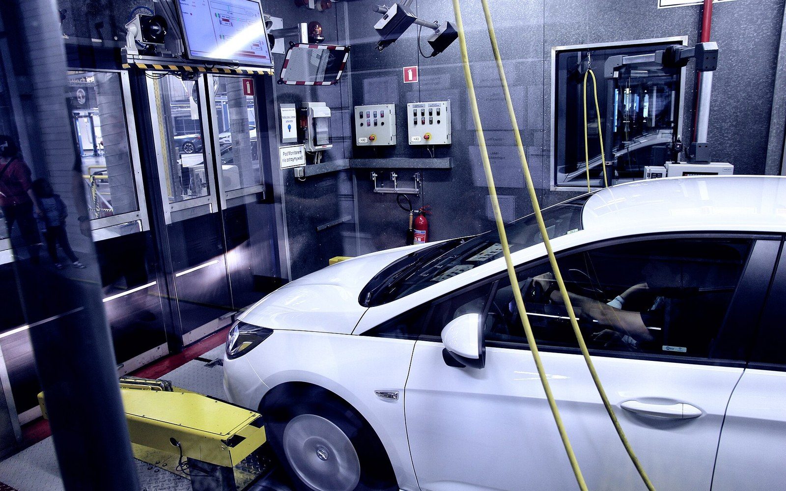 White hatchback undergoing emissions testing on a chassis dynamometer in a factory test cell